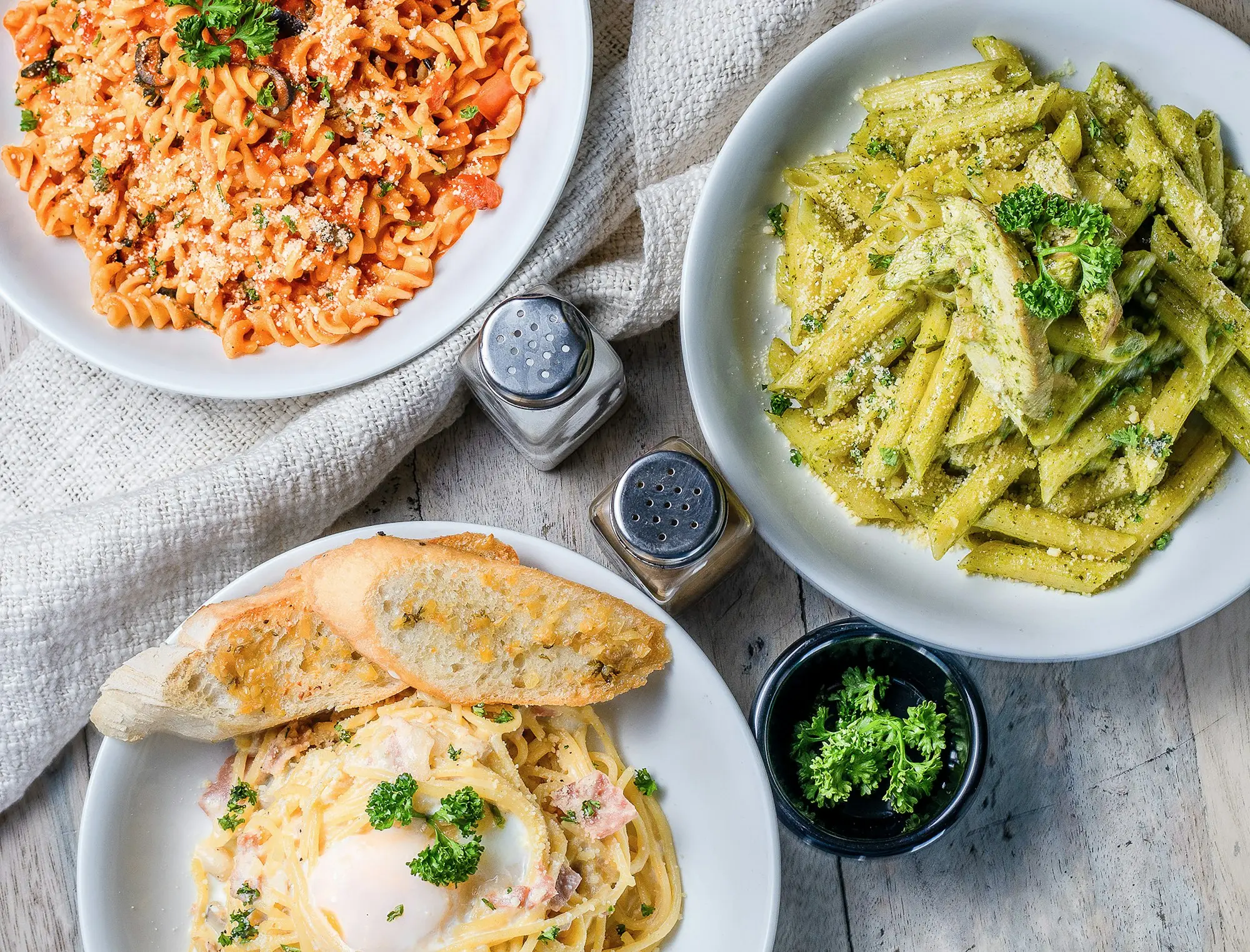 Trois assiettes de pâtes vues du dessus (fusillis à la tomate, penne au pesto et au poulet, spaghettis) avec du pain grillé