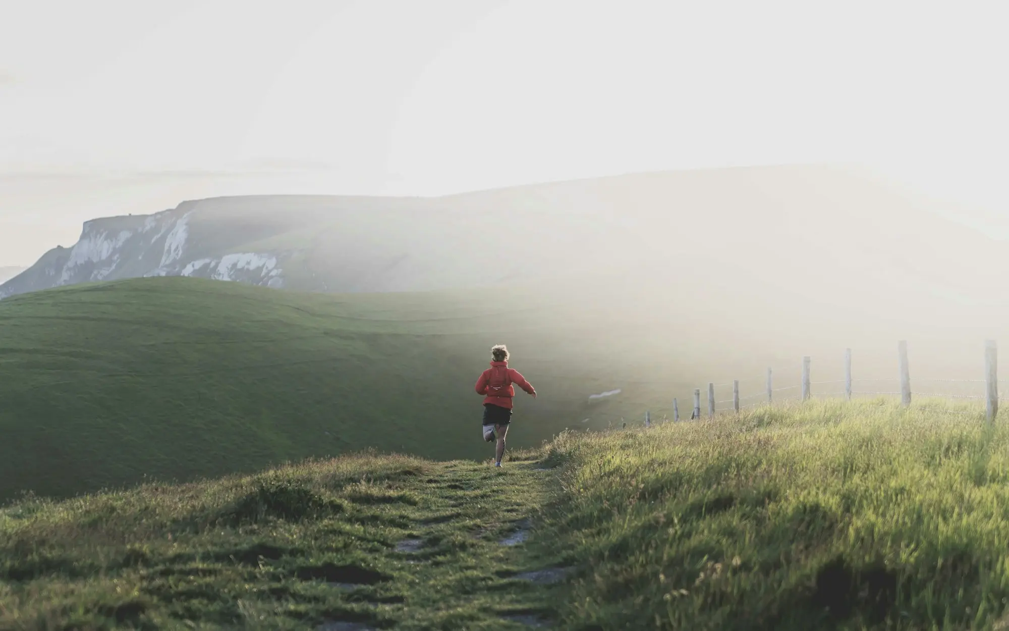 Coureur en veste rouge sur un sentier herbeux dans la brume du petit matin